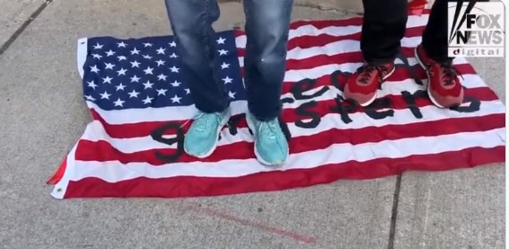 Two pro-Palestine protesters stand on an American flag with the words "Legit gangsters" written on it outside Cambridge City Hall Monday.