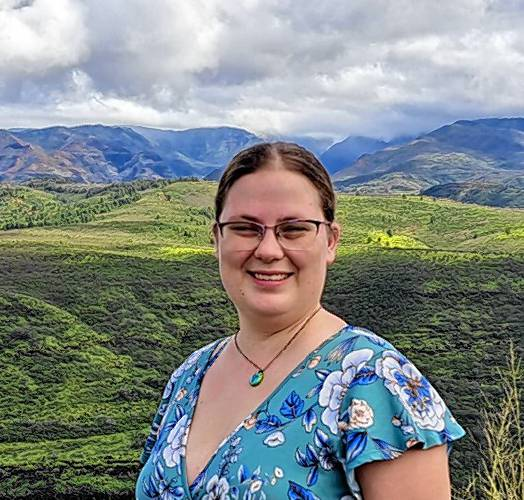A photo of Walker Powell, a woman in her 30s with a heavier build, wearing a blue short-sleeved shirt. She is pictured from the waist up, with mountains visible in the background. The photo captures her in an outdoor setting.