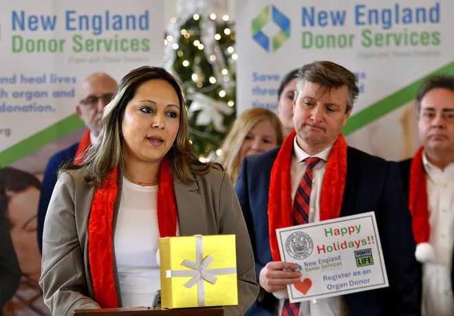 Christina Aguirre, a liver transplant recipient, speaks at a podium during an event hosted by New England Donor Services at Leominster City Hall. She is holding a gold gift box, with attendees in festive red scarves and holiday decorations in the background.