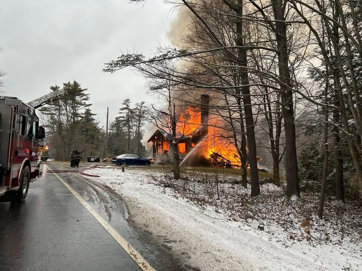A house fully engulfed in flames with fire trucks and emergency personnel present on a snowy road, trees surrounding the area. Photo Credit: Ashburnham Fire Department.