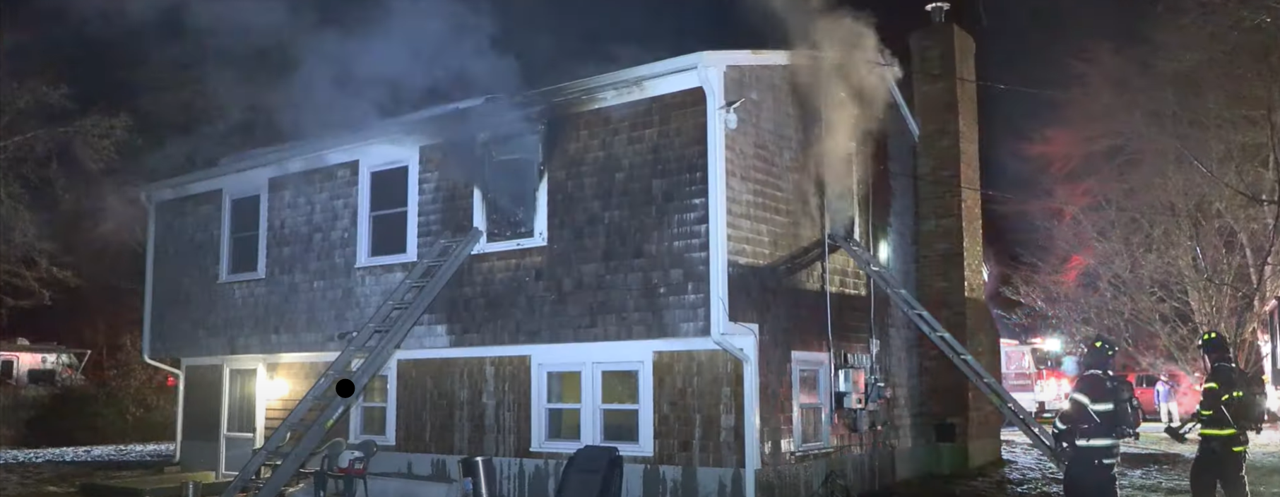 A two-story house in West Yarmouth with smoke rising from a second-floor window. Firefighters and ladders are visible around the home, actively responding to the fire.