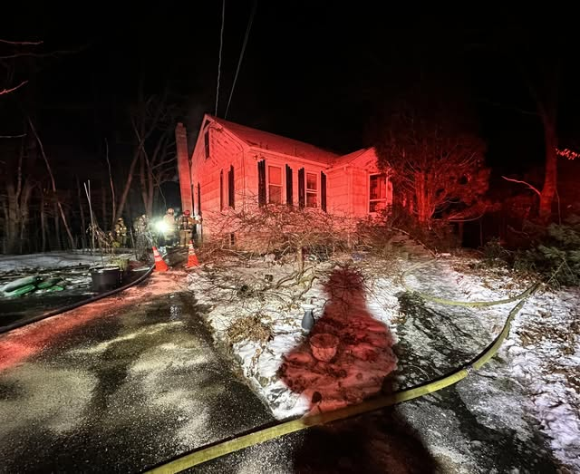 A nighttime photo of a house fire on Redemption Rock Road in Sterling, showing firefighters and emergency vehicles around a small house with visible smoke, surrounded by a snowy and icy environment illuminated by emergency lights.
