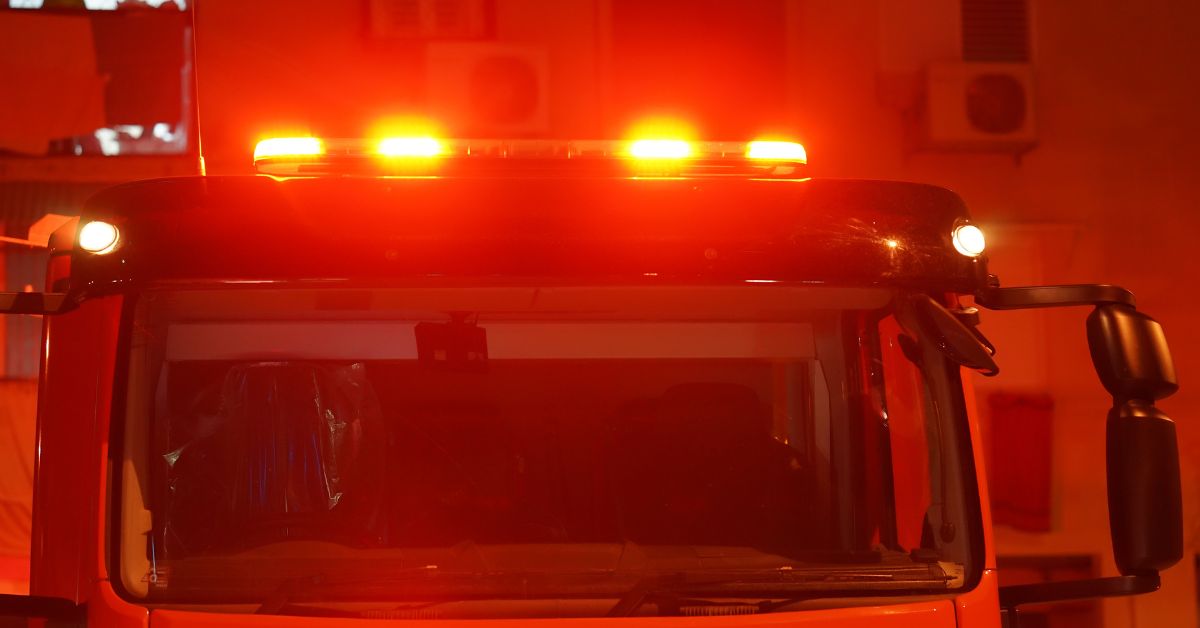 The image shows the front of a red fire engine with its emergency lights activated. The bright yellow lights on top and smaller white headlights illuminate the scene in a reddish hue. The windshield is partially visible, and the dark interior slightly obscures the driver's area. This close-up captures the intensity of a nighttime emergency response, set against a blurred background that suggests urban surroundings.