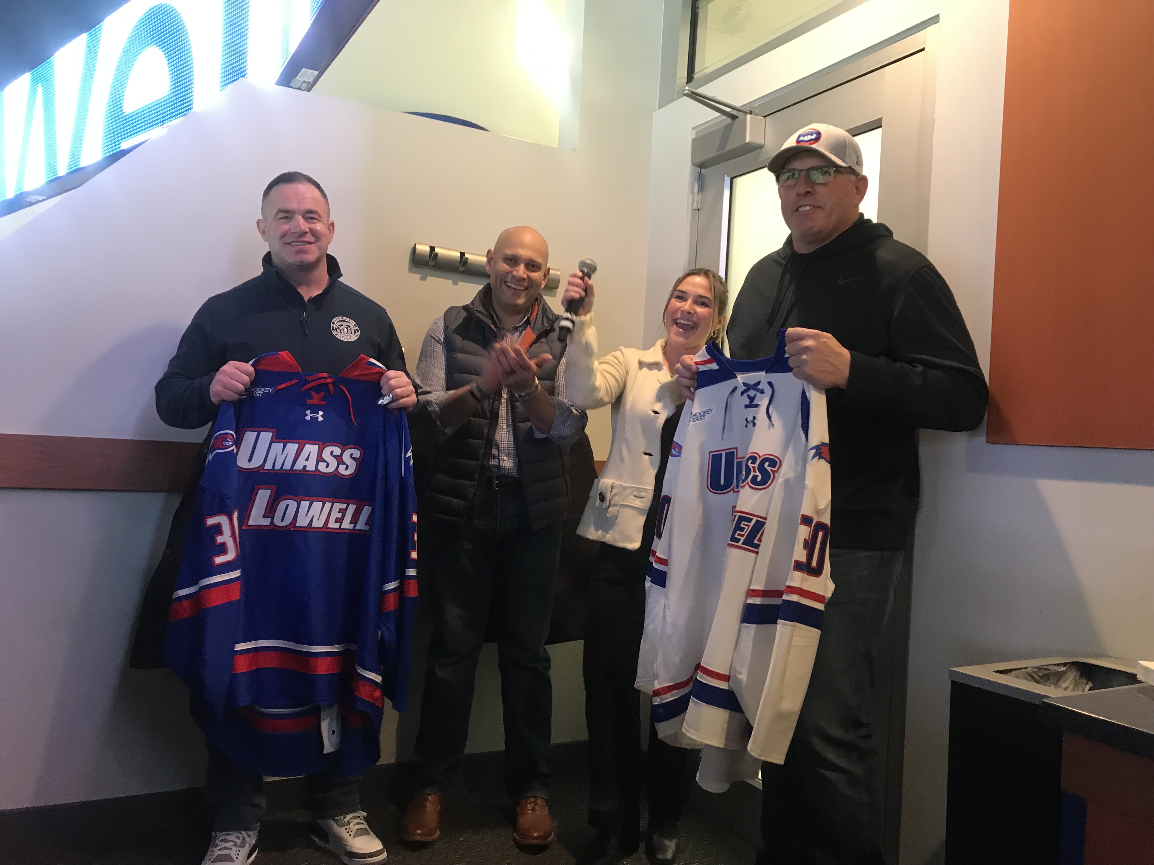 Four individuals celebrating in a room with large UMass Lowell banners. Two men on the left and right are holding UMass Lowell hockey jerseys, displaying them proudly. A woman in the center is clapping and smiling broadly, standing between a man on her left who is bald and another on her right in a baseball cap. All appear joyful in a festive atmosphere.