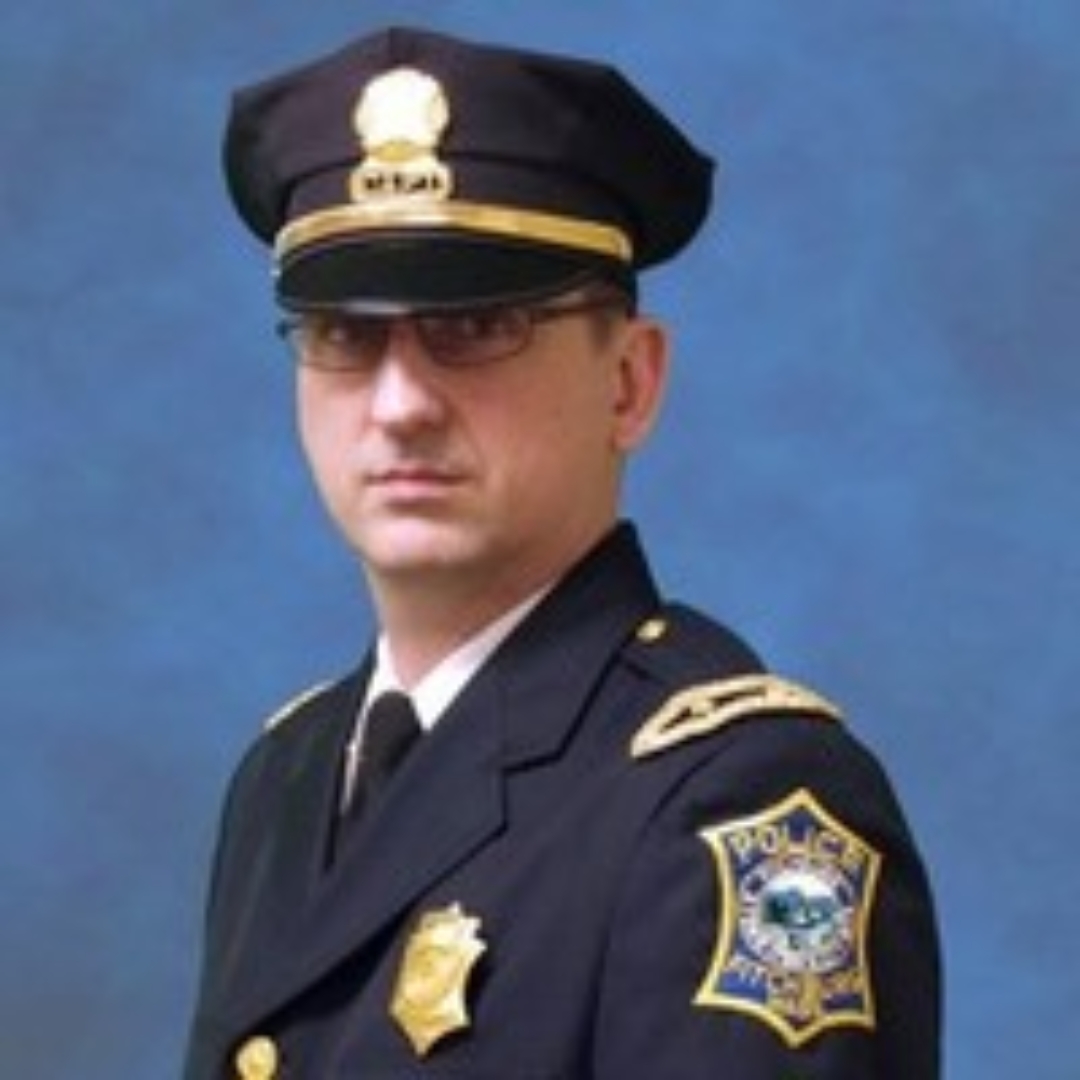 Portrait of a police officer in uniform. He is wearing a dark navy blue uniform with a police badge and insignia on the jacket. His hat features a gold-colored badge and he is looking seriously at the camera. The background is a simple blue backdrop