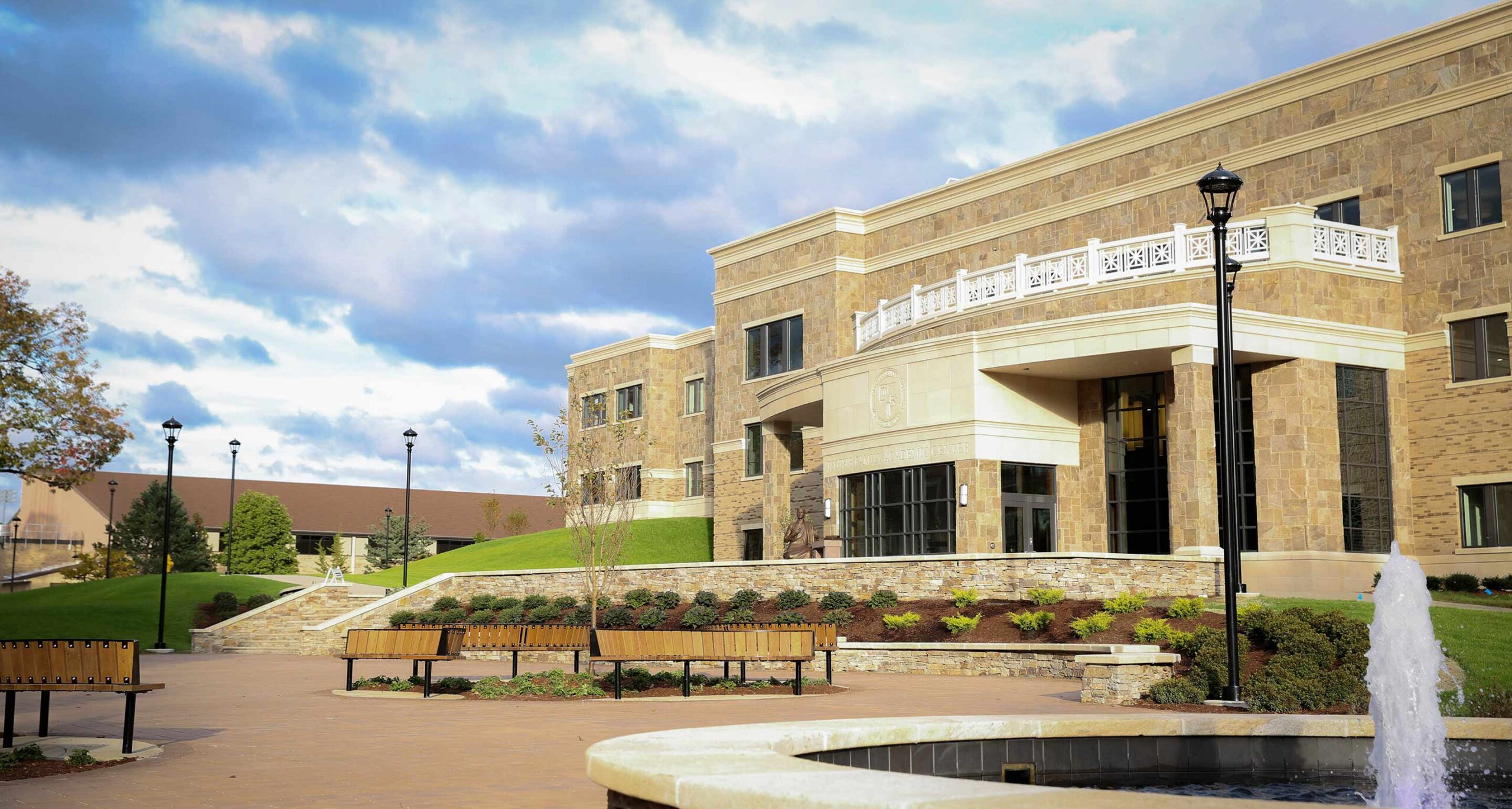 A scenic view of Assumption University's campus, highlighting the Tsotsis Family Academic Center, a stone building with large windows and a balcony. The image includes a foreground with benches, landscaped greenery, and a fountain under a partly cloudy sky.