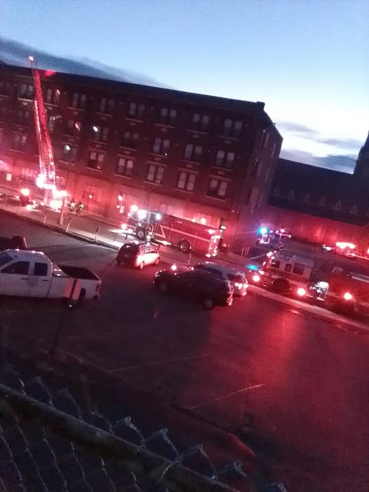 An early morning scene showing emergency vehicles with flashing lights responding to a fire at the Hotel Raymond in Fitchburg. Firefighters are using a ladder truck to access the upper floors of the brick building.