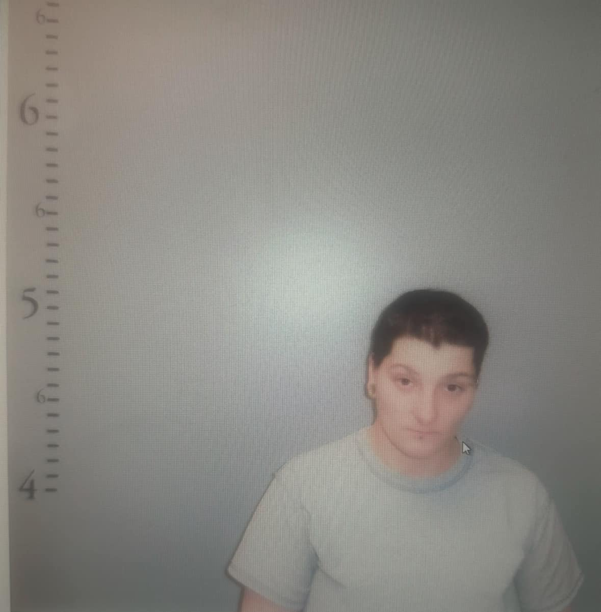 A photograph showing a person standing in front of a height chart at a police station, appearing to be around 5 feet 5 inches tall, wearing a plain white t-shirt.