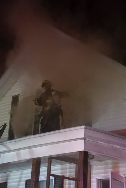 A firefighter stands on the roof of a porch, surrounded by thick smoke billowing from the upper floor of a residential structure at night.
