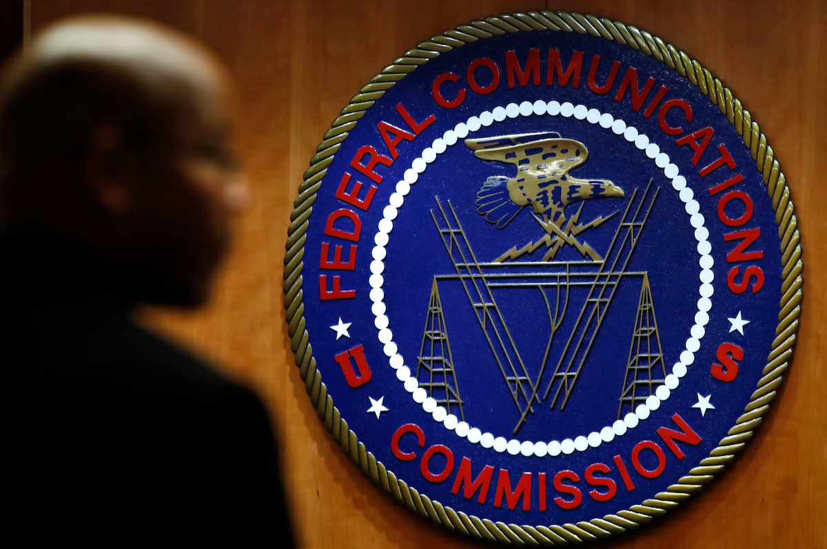 The Federal Communications Commission (FCC) seal displayed on a wooden wall, featuring an eagle, lightning bolts, and broadcasting towers, with the words "Federal Communications Commission" encircling the emblem. A blurred silhouette of a person is visible in the foreground.