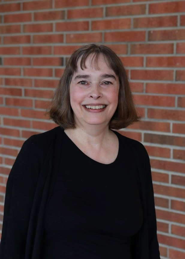 Portrait of a middle-aged Caucasian woman smiling, standing against a red brick wall background. She has shoulder-length brown hair, is wearing a black top with a draped neckline, and appears cheerful and approachable.