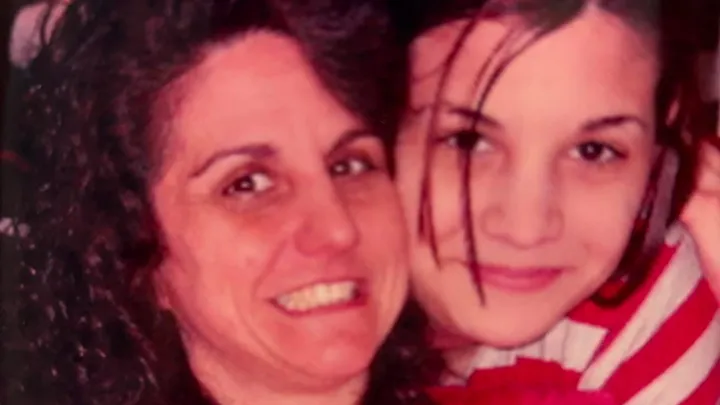 A close-up photo of two individuals, likely a mother and her daughter. The mother, on the left, has curly dark hair, a wide smile, and is wearing a dark top. The daughter, on the right, is younger with straight hair partially obscuring her face, and is wearing a striped red and white scarf. Both are posing closely, looking happy and affectionate.