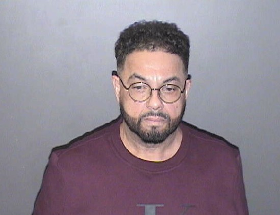 A mugshot of a middle-aged man with short curly hair and glasses. He wears a maroon T-shirt and looks directly at the camera with a serious expression. His face shows a light beard and mustache.