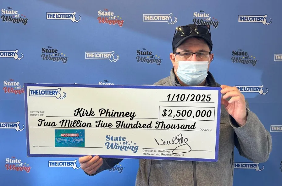 A man wearing a face mask holds a large promotional check from the Massachusetts State Lottery. The check displays his name, Kirk Phinney, and the amount of $2.5 million, dated 1/10/2025, for the “$2,500,000 Merry & Bright” scratch ticket game.