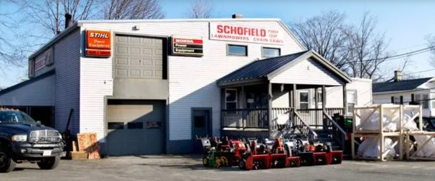 Exterior view of Schofield Power Equipment, a two-story commercial building with signage for STIHL products. The front area is lined with various models of lawnmowers and chainsaws on display. A pickup truck is parked to the left side of the building.