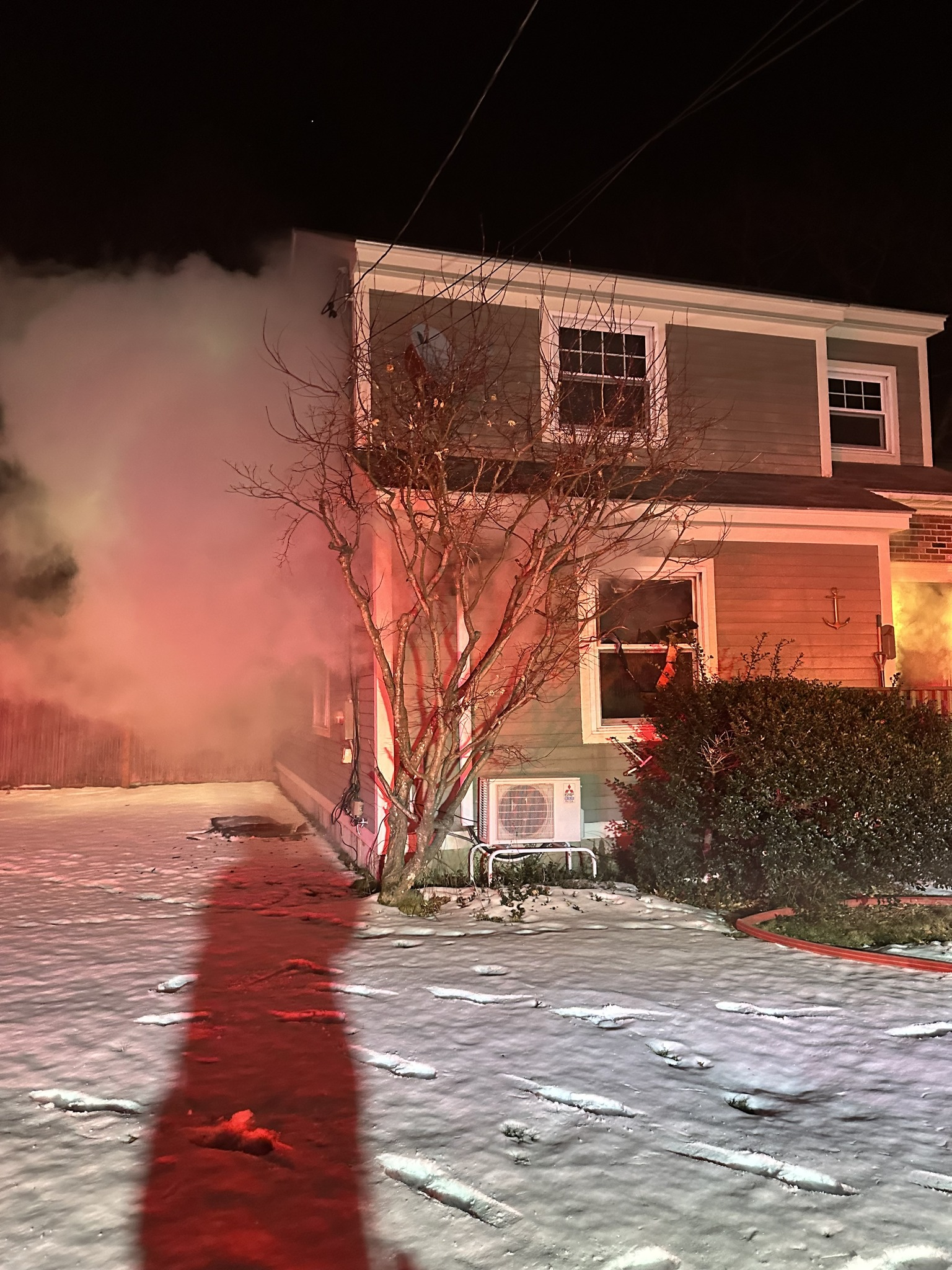 A nighttime scene of a house fire. Smoke billows from the side of a two-story home illuminated by emergency lighting, casting a red glow. The ground is covered with snow, reflecting the red light and showing footprints leading towards the house. A bare tree and a bush are visible in the foreground, also tinted red by the lighting.