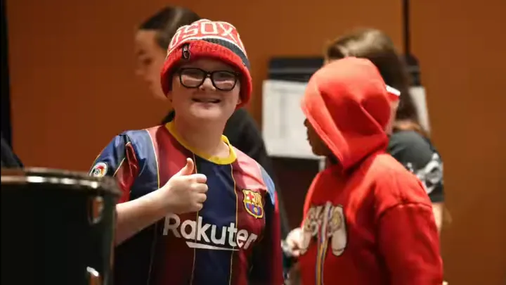 A close-up of a young boy with red hair wearing black-rimmed glasses and smiling. The background is softly blurred, suggesting an indoor setting.