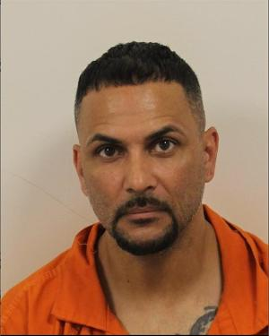 Mugshot of a middle-aged man with short black hair, a trimmed beard, and mustache. He has a stern expression and is wearing an orange jail uniform. Visible on his neck is a tattoo peeking out from under the collar