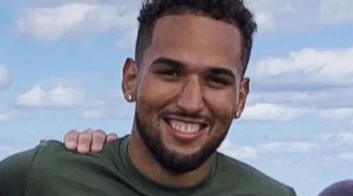 A young man with short curly hair and a beard smiles at the camera while wearing a green athletic shirt. The background features a bright blue sky with scattered clouds. His arm is around someone partially visible beside him.