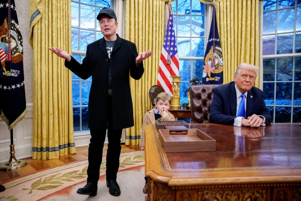 Elon Musk, wearing a black coat and cap, gestures while speaking in the Oval Office. Former President Donald Trump sits at the Resolute Desk, smiling. A young child, identified as Musk’s son X, is also present, sitting near the desk with his hand near his face. The room is adorned with golden curtains, the presidential seal, and American flags.