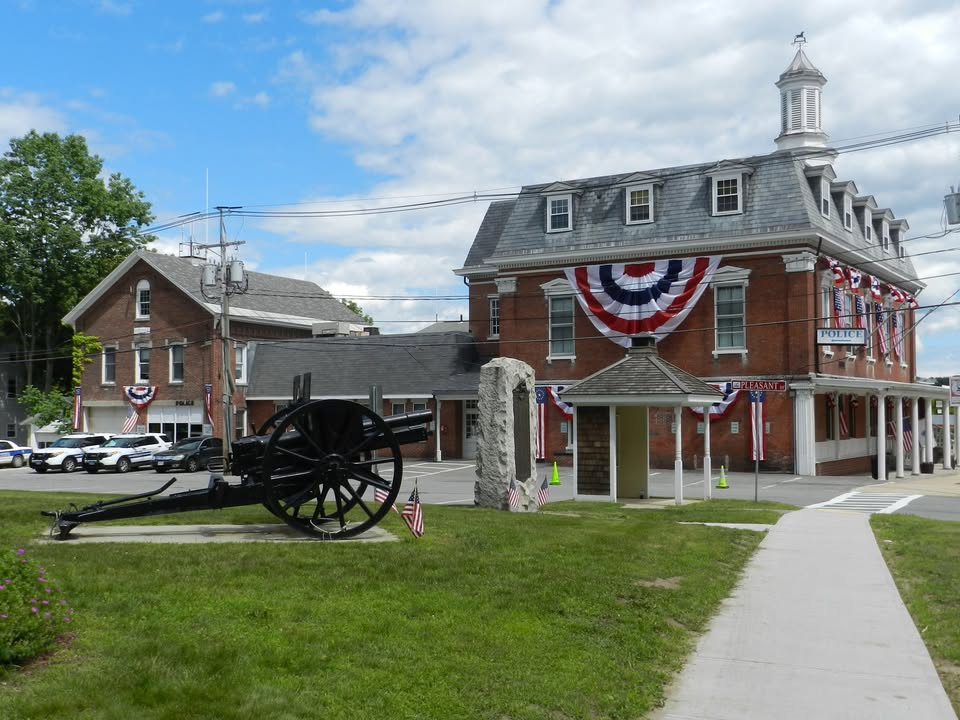 A historic red-brick building adorned with patriotic bunting, identified as Winchendon Town Hall, with a small cupola on the roof. In front of the building is a cannon monument, a stone marker, and a small gazebo-like structure. Police vehicles are parked nearby, and American flags decorate the scene.