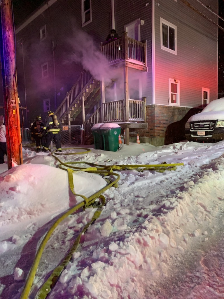 Firefighters respond to a residential fire in the Oak Hill district of Fitchburg, Massachusetts, during the early morning hours. Thick smoke is seen billowing from the first floor of a multi-story building as fire crews work in snowy conditions with hoses laid out across the icy ground.