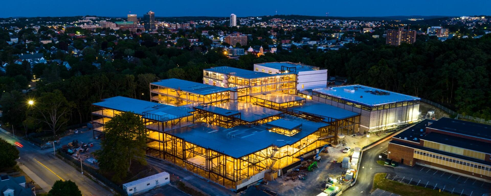 An aerial nighttime image of the new Doherty Memorial High School under construction in Worcester, Massachusetts. The building's steel framework and interior lights contrast with the surrounding cityscape in the background. (Photo credit: Fontaine Bros.)