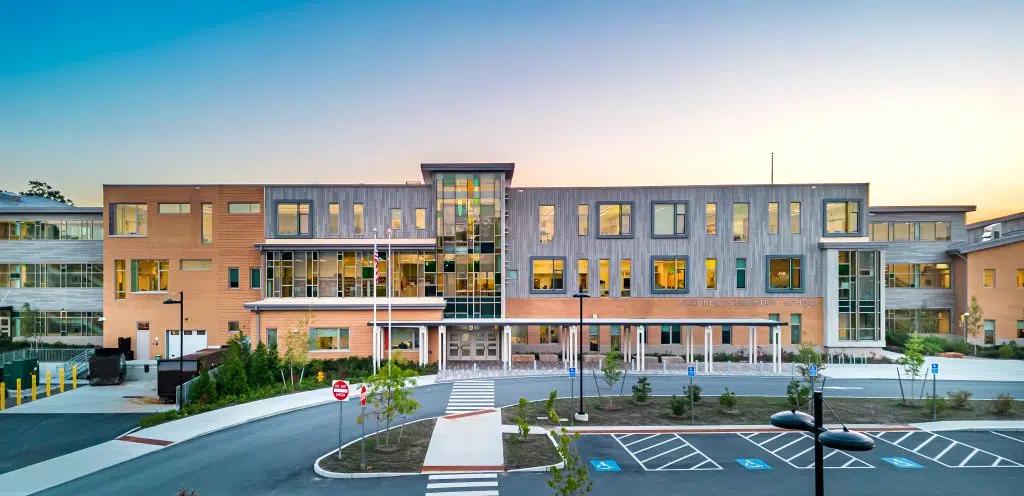 A multi-story school building with a contemporary wood-like facade and large windows, viewed at sunset, with a parking lot and pedestrian crossing in the foreground.