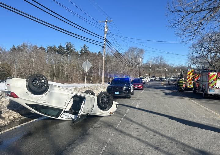 A white sedan is overturned on its roof on a two-lane road, with its driver-side door open. Police vehicles with flashing blue lights and fire trucks are present at the scene. Emergency responders can be seen working in the background. The road is lined with trees, utility poles, and a snowy roadside.