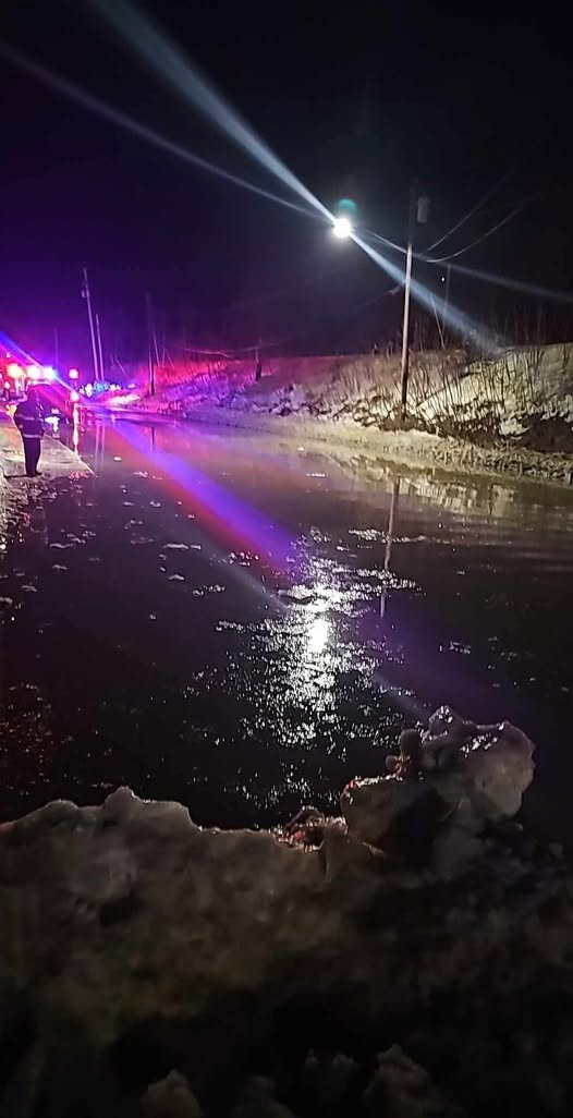 A nighttime scene of a flooded roadway in Fitchburg, with icy water covering the pavement. Emergency vehicles with flashing lights are visible in the distance, and a person stands near the edge of the water. Snowbanks line the road, illuminated by streetlights and emergency responders' lights.