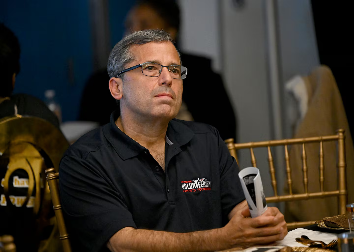 Josh Kraft, wearing a black polo shirt with the New England Patriots Foundation logo, sits attentively at an event. He appears focused, listening intently in a room with subdued lighting and a background that includes other attendees and golden event chairs.