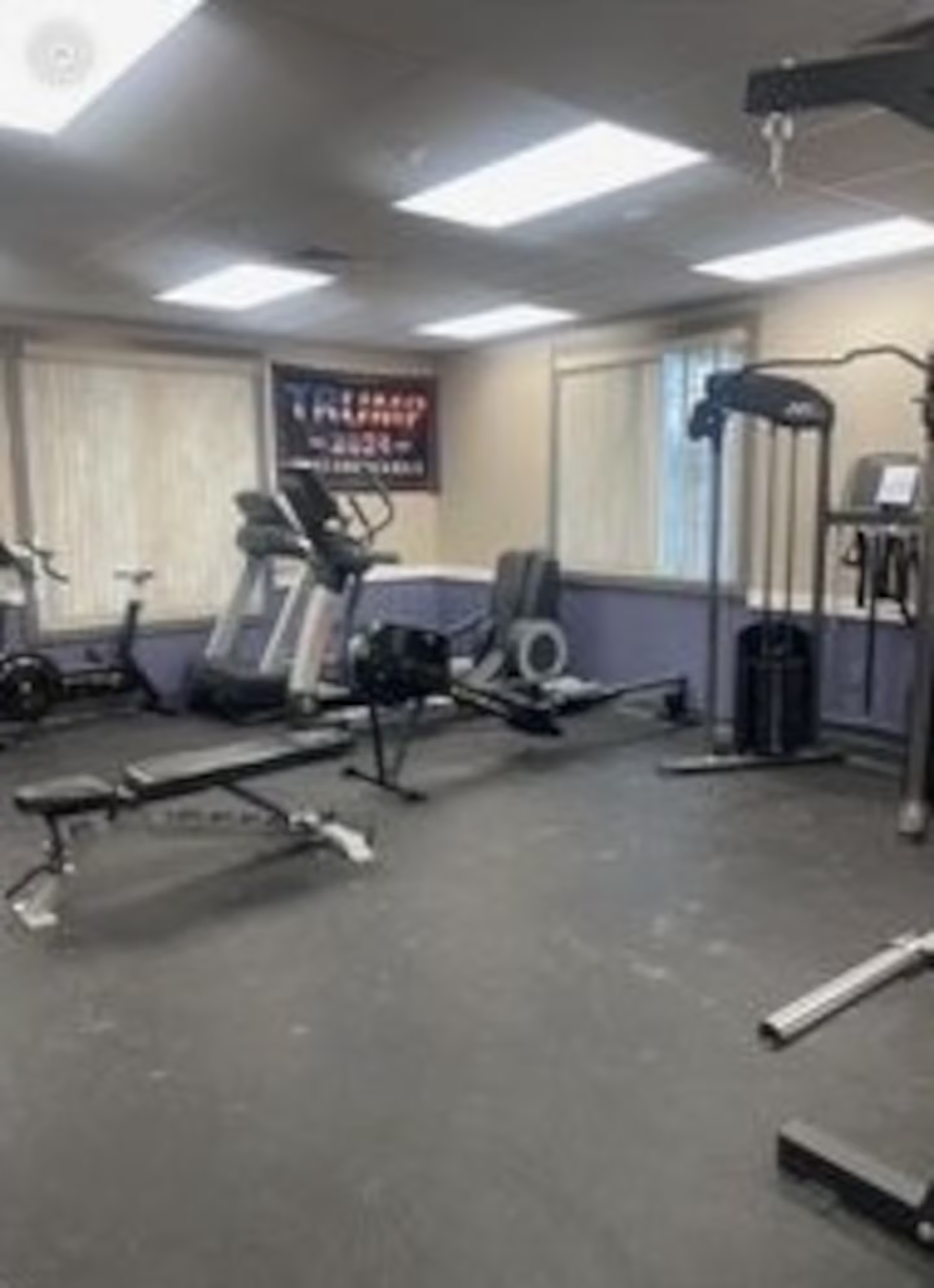 A gym with various exercise equipment, including a treadmill, weight bench, and cable machine. A Trump flag hangs on the wall in the background. The room is well-lit, with windows covered by blinds.