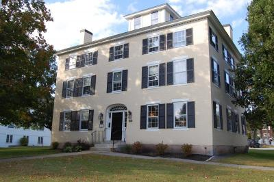 A three-story, beige historic building with black shutters and a symmetrical facade. The entrance features a decorative archway with a black door. The building is surrounded by a well-maintained lawn with small bushes near the foundation. Large trees frame the property, and a bright blue sky with scattered clouds is visible in the background.