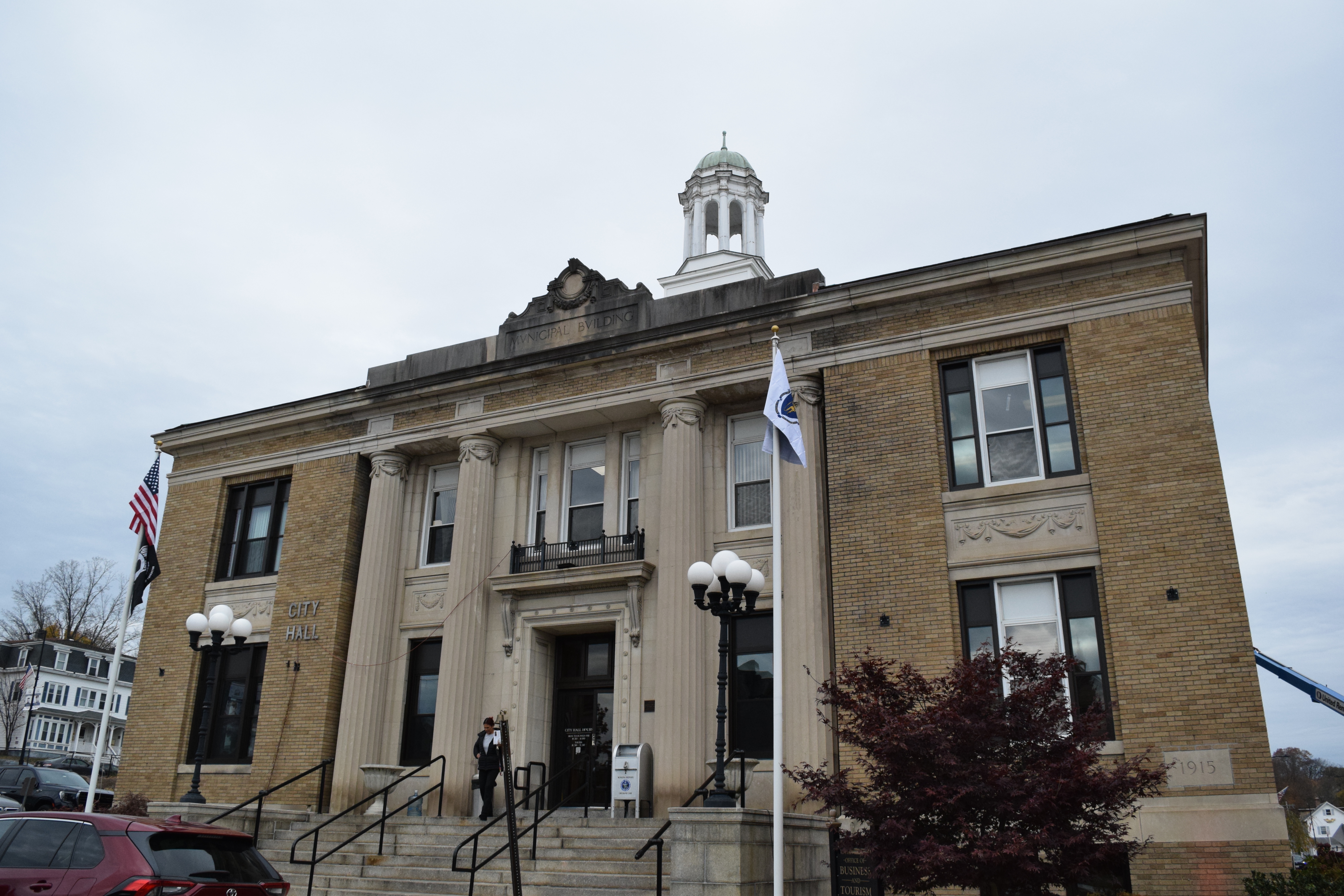 Front view of Leominster City Hall, a historic brick building with large columns, flags, and a cupola under an overcast sky.