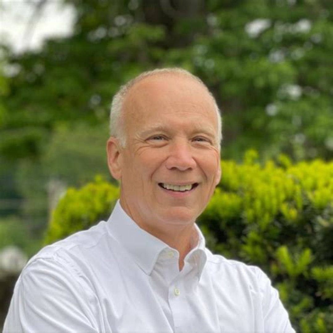 A middle-aged man with short-cropped white hair smiles while wearing a white button-down shirt. He is outdoors with lush green trees and bushes in the background.