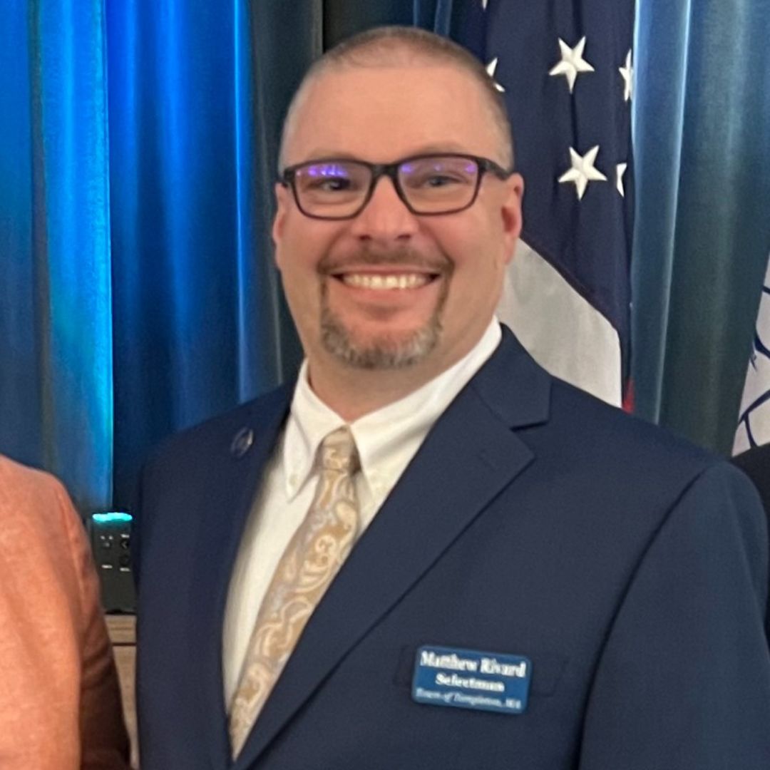 A smiling Matthew Rivard wearing a navy blue suit, white shirt, and patterned tie, standing in front of an American flag and blue curtains. He has glasses and a neatly trimmed beard, with a name tag that reads "Matthew Rivard, Selectman, Town of Templeton, MA."