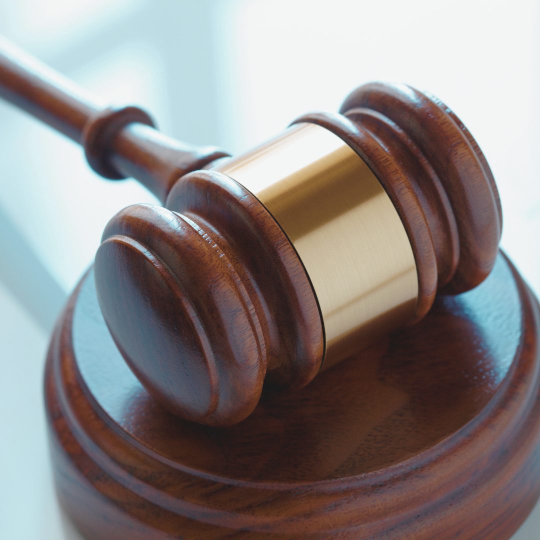 The image shows a close-up of a wooden gavel with a brass band on the handle, resting on its base. The background is blurred, suggesting a courtroom or legal setting.