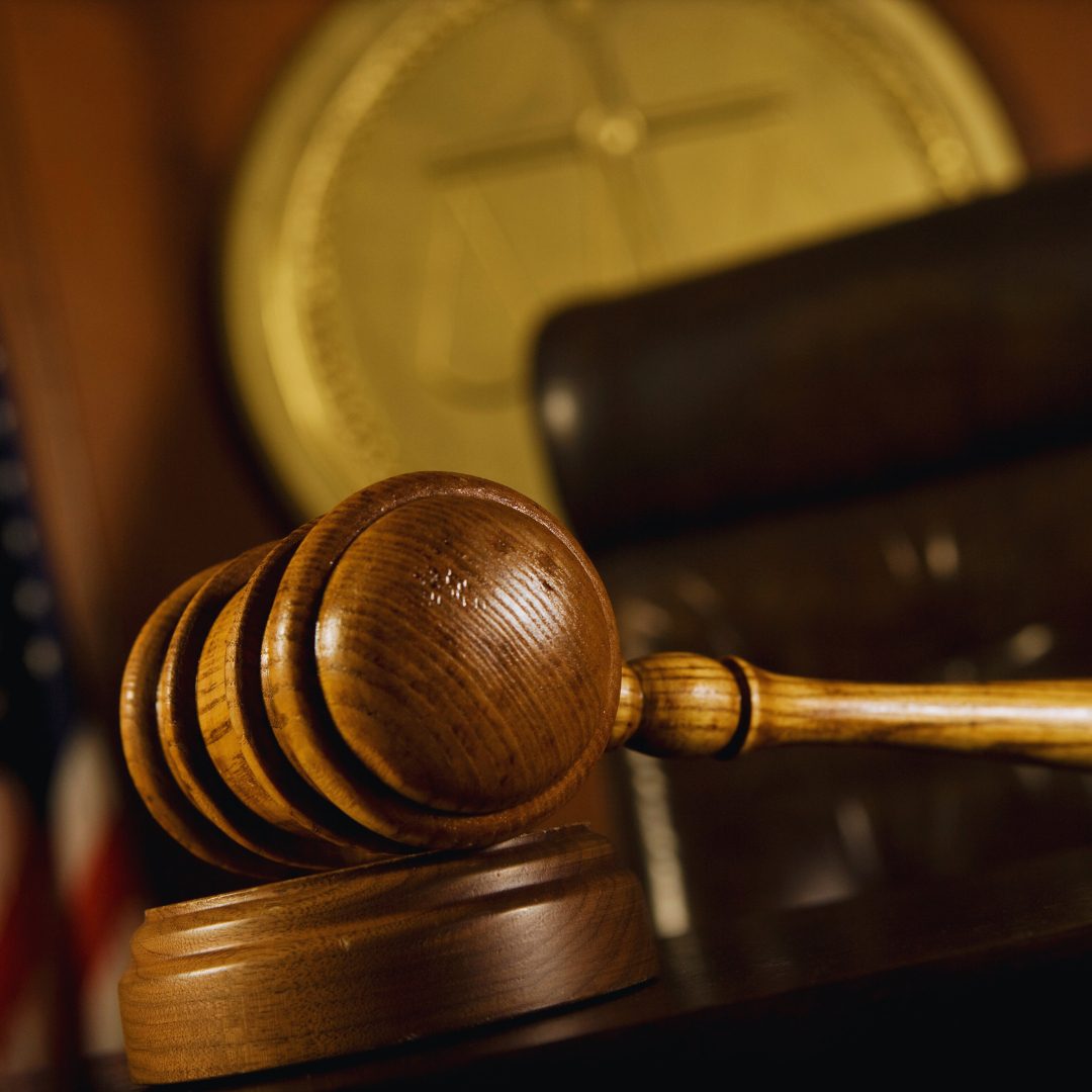 The image shows a close-up of a wooden gavel with a smooth handle and multiple grooves on the head, resting on its base. In the background, there is a blurred representation of a golden scale of justice.