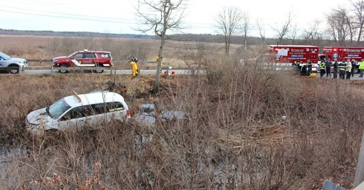 A white van partially submerged in marshy water beside a roadway, with emergency vehicles and responders from Sudbury and Wayland visible in the background.