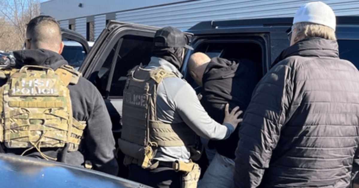 ICE agents wearing tactical gear arrest a suspect in Massachusetts, placing them into a vehicle during a large-scale enforcement operation that resulted in over 370 arrests. The agents are dressed in protective vests, with one displaying "HSI POLICE" on the back.