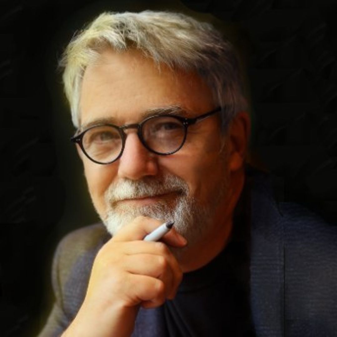 A portrait of Peter H. Reynolds, a bestselling author and illustrator, wearing black-framed glasses and a dark blazer. He has white hair and a beard, and he is resting his chin on his hand while holding a pen. The background is dark and softly blurred.