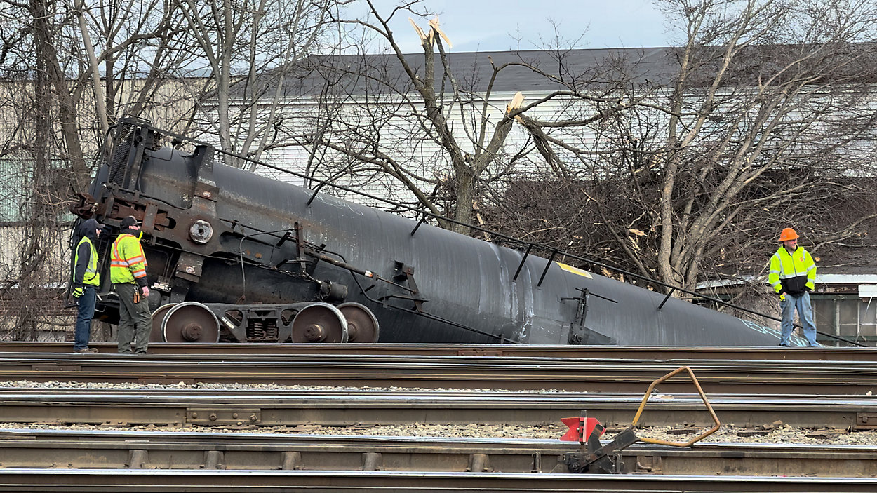 Emergency crews inspect a black tanker car that derailed in Worcester on Sunday. The rail car is tilted off the tracks, with responders wearing high-visibility gear assessing the damage. Trees with broken branches and industrial buildings are visible in the background. (Photo by Spectrum News 1/Devin Bates)