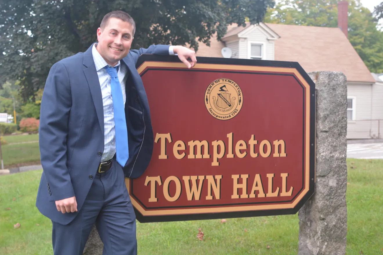 A man in a navy blue suit, light blue tie, and checkered dress shirt leans on a maroon and gold "Templeton Town Hall" sign with one hand while smiling at the camera. A house with beige siding and a satellite dish is visible in the background, along with green trees and grass.