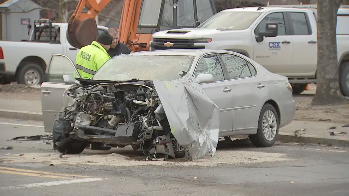 A heavily damaged silver sedan with its front end completely destroyed is seen on the road after a collision. A police officer in a neon yellow jacket marked "POLICE" and another individual are inspecting the wreckage. In the background, a CSX service truck and construction equipment are visible.