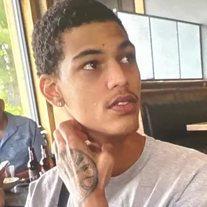 A young man with short, curly black hair and a light brown complexion is seen wearing a light gray t-shirt. He has a tattoo on his hand and is touching his neck while gazing off to the side. The background shows a restaurant setting with booths, large windows, and bottles on the table.