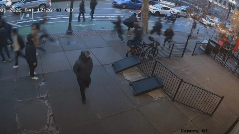 A surveillance camera image captures a group of people running on a sidewalk outside a restaurant in Back Bay. Some individuals appear to be looking back while others move quickly in different directions. A police vehicle is visible in the background on the street.