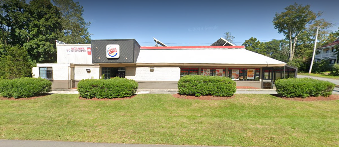 A Burger King restaurant with a white roof and black signage sits in a suburban area surrounded by green bushes and grass. Large windows display promotional posters for menu items. A sign on the building reads "We're Open Treat Yourself." Trees and a residential house are visible in the background.