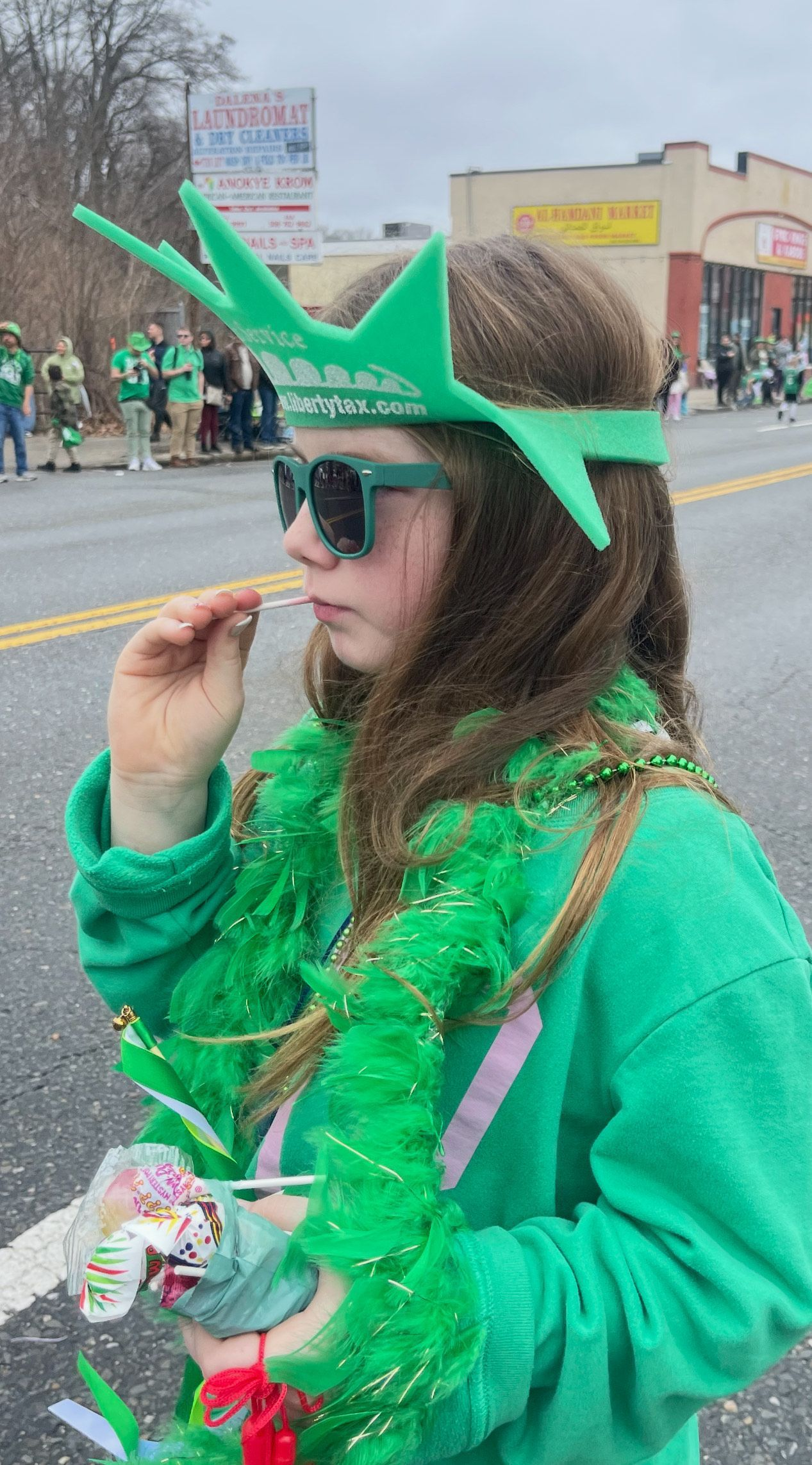 A girl watches a parade on Park Avenue. She wears a green hoodie, a feather boa, beaded necklaces, green sunglasses, and a foam Statue of Liberty-style crown with "Liberty Tax" printed on it. She holds a bag of candy and a lollipop while observing the parade. In the background, parade-goers dressed in green line the street, with storefronts and signs visible.