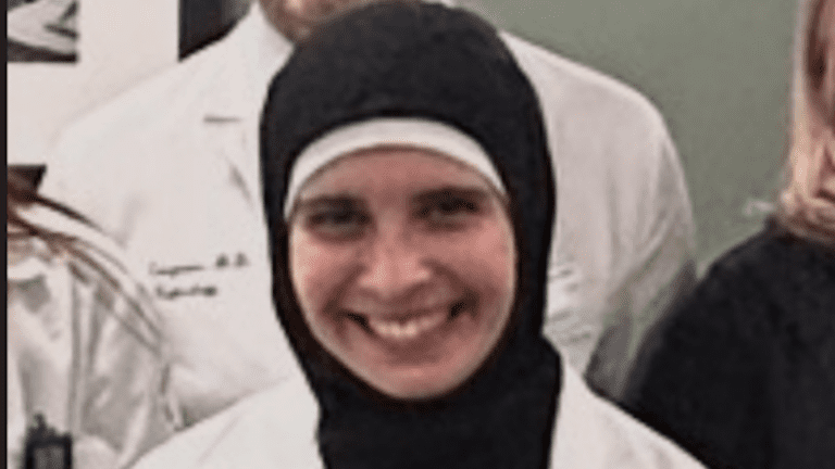 Dr. Rasha Alawieh smiling, wearing a black hijab and a white medical coat, standing among colleagues in a professional group setting.