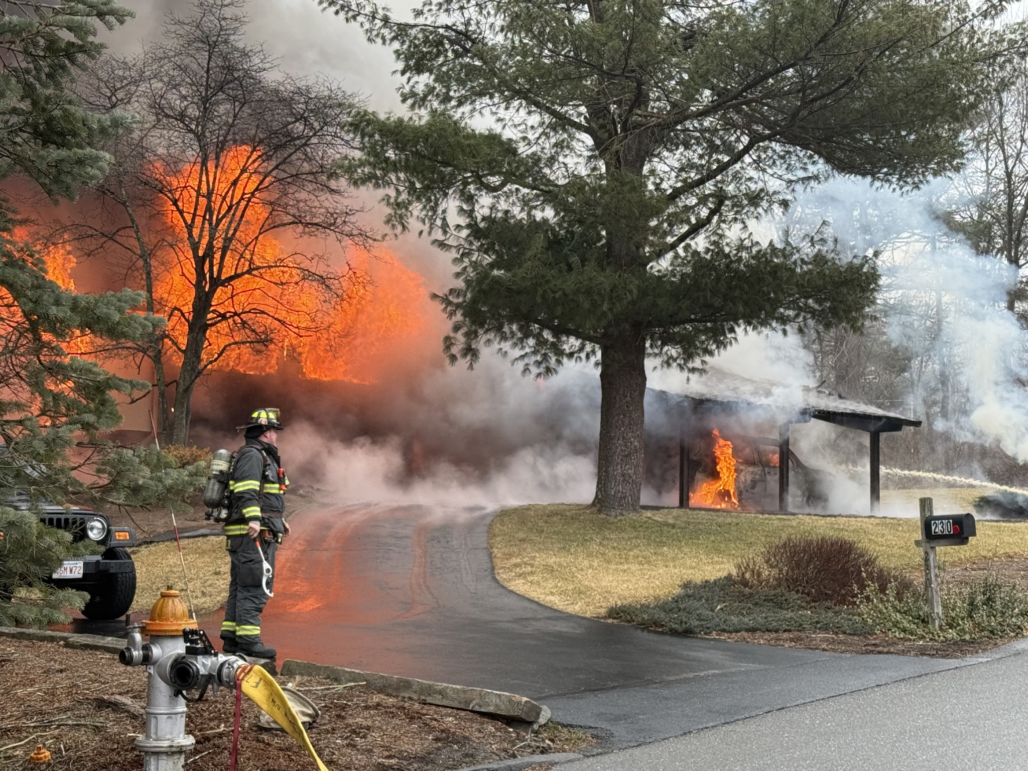 A firefighter stands near a hydrant as flames engulf a single-story home, with thick smoke billowing into the sky. A vehicle under the carport is also on fire.